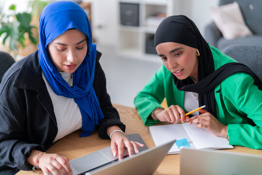 Two women in hijabs discuss work while sitting at a desk with laptops in front of them. The bright and modern office setting enhances their focused collaboration.