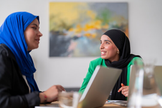 Two women in hijabs discuss work while sitting at a desk with laptops in front of them. The bright and modern office setting enhances their focused collaboration.