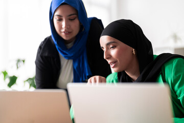 Two women in hijabs discuss their work while using laptops in a modern, bright office. The image captures a collaborative and professional atmosphere.