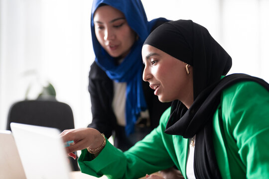 Two women in hijabs discuss their work while using laptops in a modern, bright office. The image captures a collaborative and professional atmosphere.