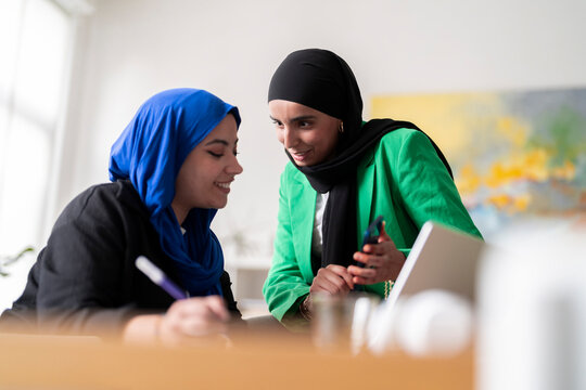 Two women in hijabs discuss and review work while one writes and the other holds a phone in a modern office. The image captures a collaborative and professional atmosphere.