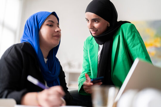 Two women in hijabs discuss and review work while one writes and the other holds a phone in a modern office. The image captures a collaborative and professional atmosphere.