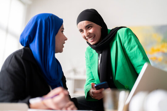 Two women in hijabs discuss and review work while one writes and the other holds a phone in a modern office. The image captures a collaborative and professional atmosphere.