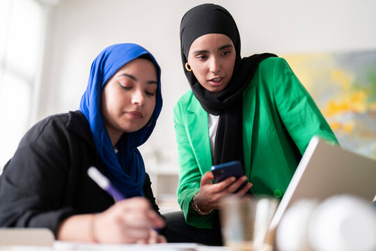 Two women in hijabs discuss and review work while one writes and the other holds a phone in a modern office. The image captures a collaborative and professional atmosphere.