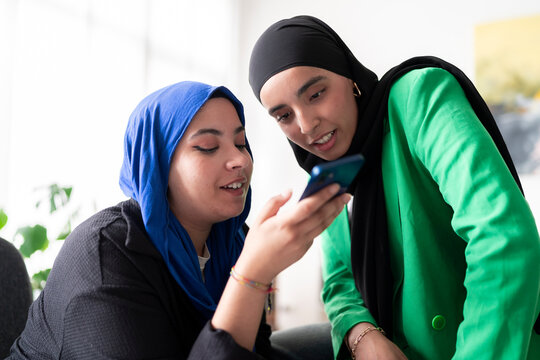 Two women in hijabs recording a voice message on mobile phone in a bright office. Their joyful expressions add to the friendly and positive work environment.