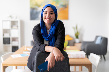 A woman in a blue hijab smiles while holding a phone and leaning on a chair in a bright office. Her relaxed and positive demeanor highlights the friendly work atmosphere.