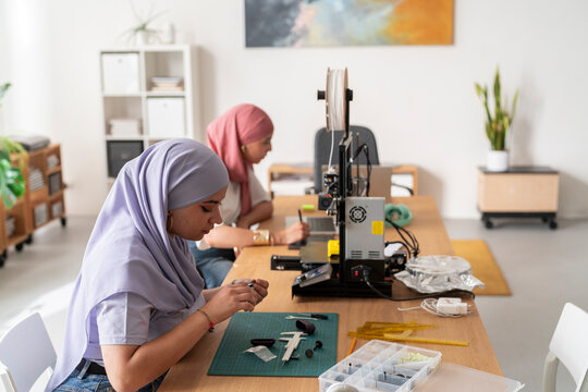 Two women in hijabs are focused on their tasks in a modern workshop. One woman is assembling a small part while the other works on a computer