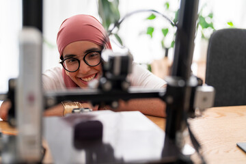 A woman in a dark red hijab smiles as she looks at the outcome of a 3D printing project. Her expression shows satisfaction and joy in the successful completion of the task.