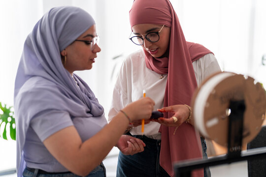 Two women in hijabs engage in a discussion about 3D printing components. They hold parts and tools, showcasing their involvement in the technical aspects of the project.