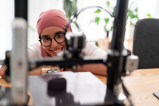 A woman in a dark red hijab smiles as she looks at the outcome of a 3D printing project. Her expression shows satisfaction and joy in the successful completion of the task.