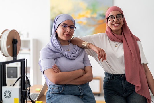 Two women in hijabs pose confidently in a 3D printing lab, surrounded by equipment. Their expressions reflect pride in their technical skills and accomplishments.