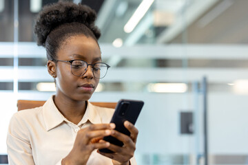 Young african american businesswoman using phone at workplace inside office. Professional woman focused on screen, wearing glasses, corporate attire. Modern business represents productivity