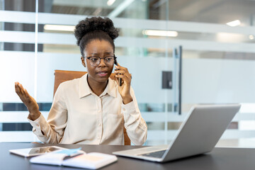 Young african american businesswoman working at office desk using laptop and phone. She appears to be having a discussion, with a serious expression. Professional workplace environment.