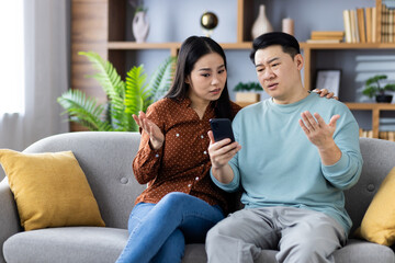 Asian couple sitting on couch looking at phone with confused expressions, discussing something important. Image shows emotional interaction, communication, relationship dynamics, technology use
