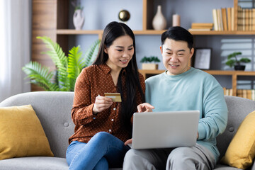 Asian couple enjoying online shopping with laptop and credit card while sitting on cozy sofa at home. Happy moment of browsing and making purchases together.