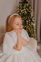 A beautiful blonde girl in a white dress has closed her eyes and is making a wish near the Christmas tree