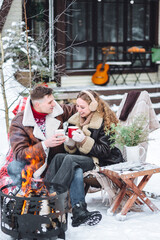 Young loving couple spending winter holidays in a countryside cottage house. Wearing warm clothes, knitted sweaters, mittens. Hot drinks. Cozy Christmas atmosphere, New Year's Eve, snowy frosty day