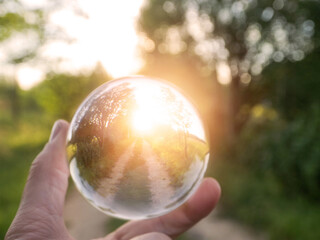 A person is holding a clear glass ball that is reflecting the sunrise over a small country road, selective focus. The scene is peaceful and serene, with the sun shining brightly in the background