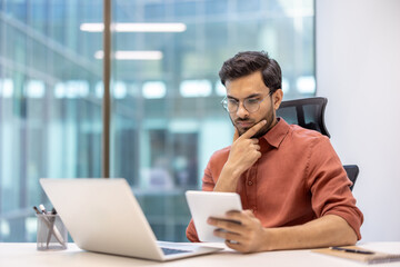 Young man in modern office using laptop and tablet. Man in casual wear concentrating on work tasks....