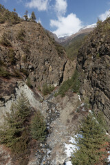 Mountain river is making its way through a narrow valley in the himalayas, surrounded by rocks, trees and snow. The sun is shining and some clouds are visible