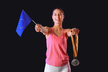 Sporty young woman with prize medals and flag of European Union on dark background
