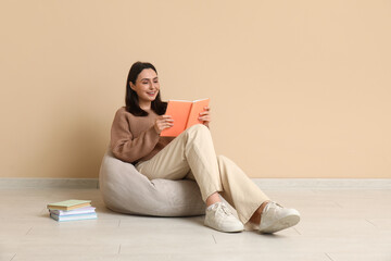 Young woman sitting on pouf and reading book near beige wall