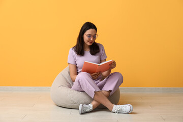 Young woman in eyeglasses sitting on pouf and reading book near orange wall