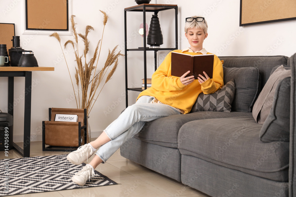 Wall mural Young woman in eyeglasses reading book and sitting on sofa in stylish office