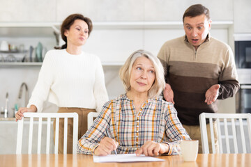 Thoughtful old woman compiling heritage document while middle-aged man and woman competing with...