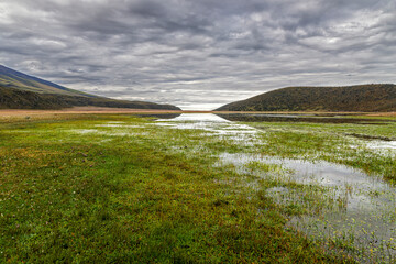 An aquatic path through the algae of a lagoon to a spot of light between two hills below a dramatic cloudy sky