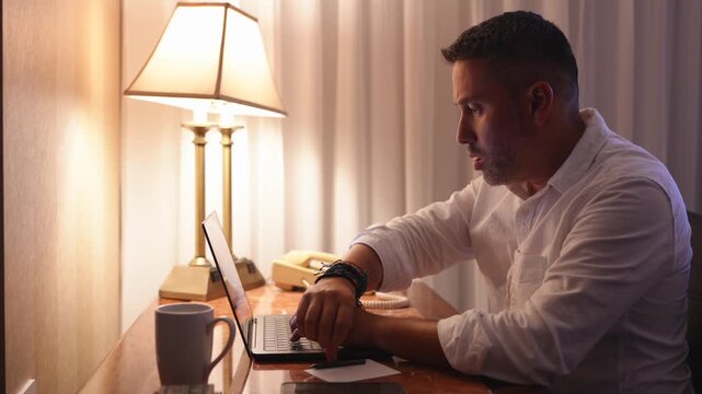 Hombre trabajando de noche frente a la computadora sentado en un escritorio en la habitaci&oacute;n de un hotel