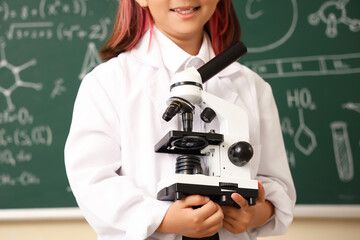 Cute little girl with microscope near chalkboard at chemistry lesson in classroom