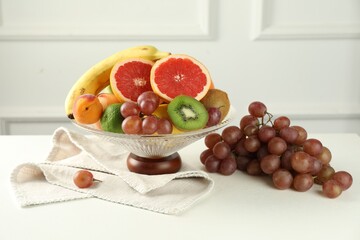 Glass vase with different fresh fruits and grapes on white table