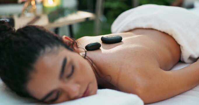 Woman, relax and back with rocks for massage, spa day or stress relief on bed at outdoor beauty resort. Closeup of young female person lying on table with masseuse in relaxation for therapy treatment