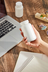 On the wooden table with laptop, glasses and notebook, hand of woman is holding a white medicine bottle without label. Creative scene for advertising, space for design