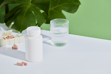 A white bottle unlabeled displayed on white table with water cup and daily medicine box. Behind are green leaves on green background. Blank space for design