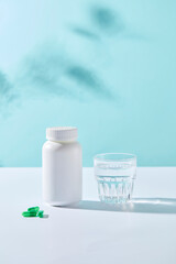 Minimal scene for advertising medicine product with blue background. Front view of white medicine bottle, glass water cup and capsules decorated. Space for design label