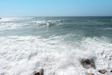 Waves crash on rocky shoreline under  clear sky during midday