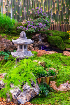 Minimal Japanese garden in home with small stone lantern and bamboo fountain and moss and flowers.

