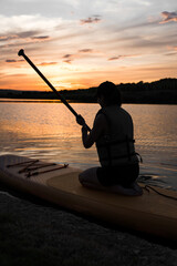 A young woman swims on a SUP board in a lake at sunset. Silhouette of a woman sitting on a SUP board at sunset. SUP surfing concept