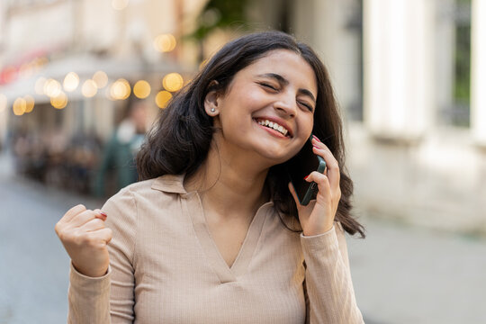 Phone call good news gossip. Happy surprised Indian woman in pleasant conversation on smartphone enjoying talking with friends outdoors. Hispanic girl walking on urban city street. Town lifestyles