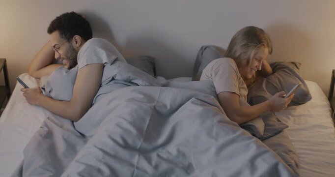 Husband and wife using smartphones ignoring each other lying on different sides of bed focused on gadgets in bedroom in evening. Marriage and modern technology concept.
