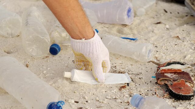 Beach cleanup effort, hand picking up plastic toothpaste tube in sandy beach, water bottles and discarded footwear 