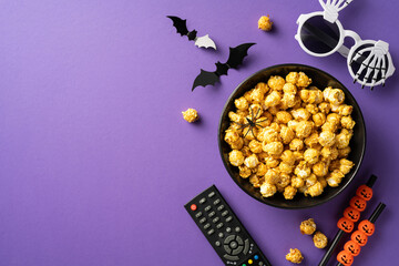 Bowl of caramel popcorn surrounded by Halloween-themed decorations like paper bats, fake spider, and  skeleton glasses on a purple background