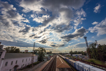 Trains line the platform beneath an expansive sky painted with dramatic clouds during the golden hour of late afternoon.