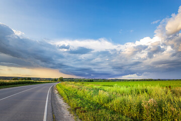A winding asphalt road leads through a verdant field of tall grasses, bordered by a line of trees, with a dramatic sky filled with clouds and streaks of sunshine overhead.