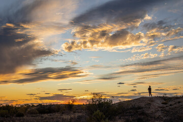 A woman standing on a mound of rock and silhouetted by the setting sun against a glorious and colorful sunset sky with cumulus clouds in the remote Currawinya National Park in Queensland, Australia.