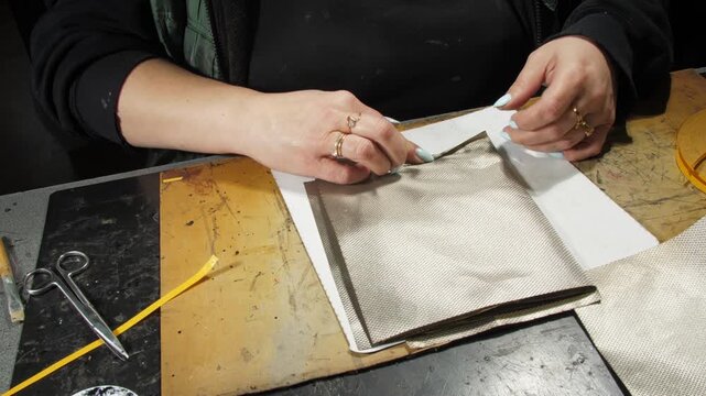 Seamstress hands roll up lining fabric combined details at desk closeup. Woman makes metallic material casing part in sewing workshop