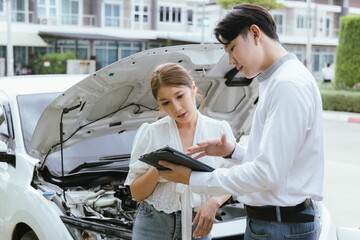 A young Caucasian man and an Asian woman inspect a damaged car after an auto accident. The insurance agent examines the vehicle, writing a report while gathering evidence for the claim investigation a