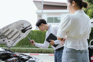 A young Caucasian man and an Asian woman inspect a damaged car after an auto accident. The...